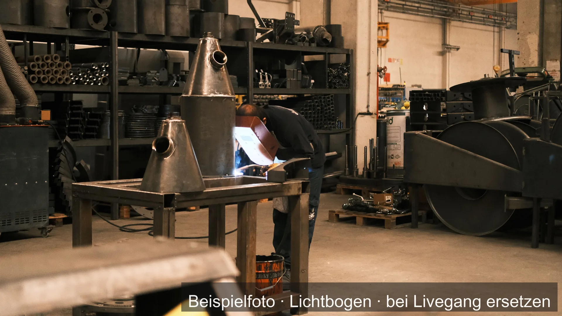 Ein Mann schweißt in einer Werkstatt an kegelförmigen Stahlteilen, dahinter Regale mit Material.