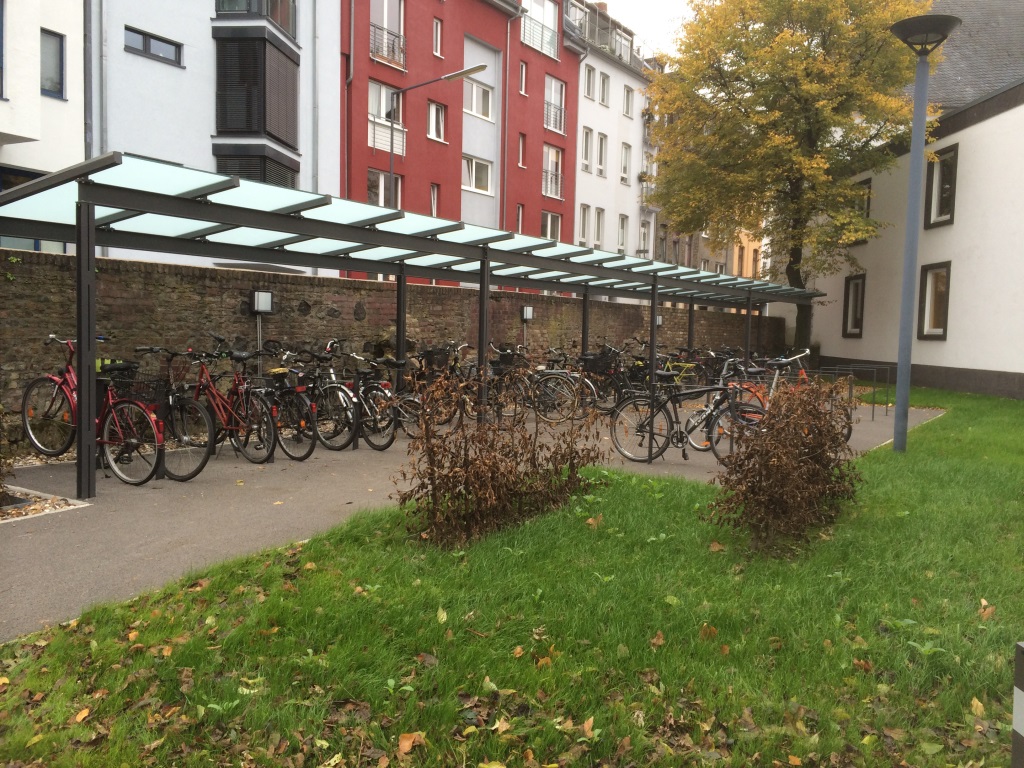 Lange Fahrradüberdachung aus dunklen Stahlstützen und -trägern mit aufgesetzten Glasscheiben vor Wohnhäusern, darunter abgestellte Fahrräder.