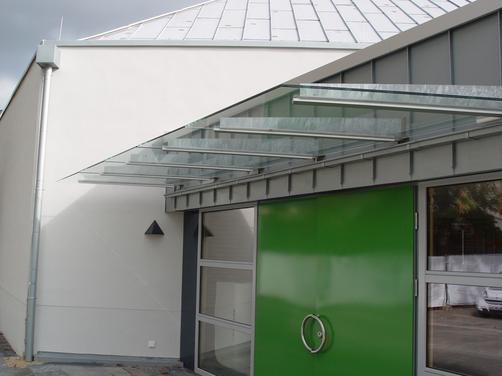Glasvordach aus verzinkten Stahlschwertern mit unterseitig befestigten Glasscheiben über einer grünen Doppeltür an einer hellen Hallenfassade.