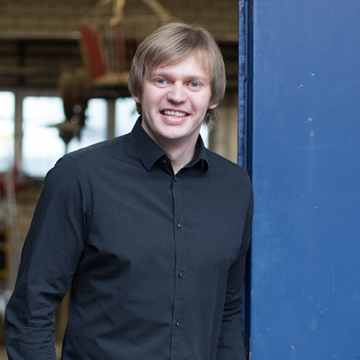 Harry Riesen, Projektleiter, lächelnd im schwarzen Hemd neben einer blau beschichteten Stahlstütze, dahinter eine Werkhalle.