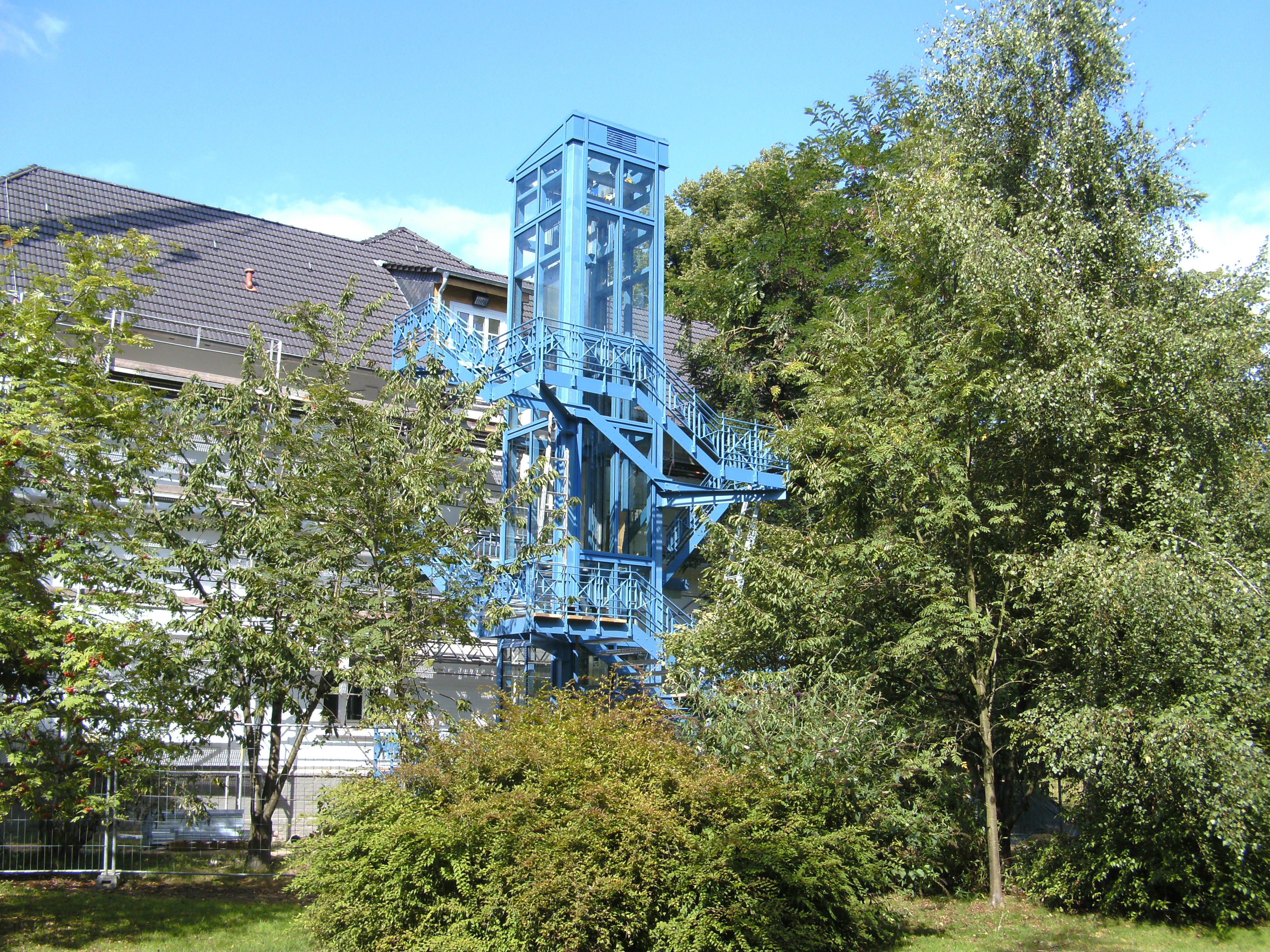 Außentreppe für den Landschaftsverband Langenfeld, mehrläufig mit Podesten und Stabgeländer, an einem hellblau beschichteten Turm in Stahlrahmen-Bauweise zwischen Bäumen.