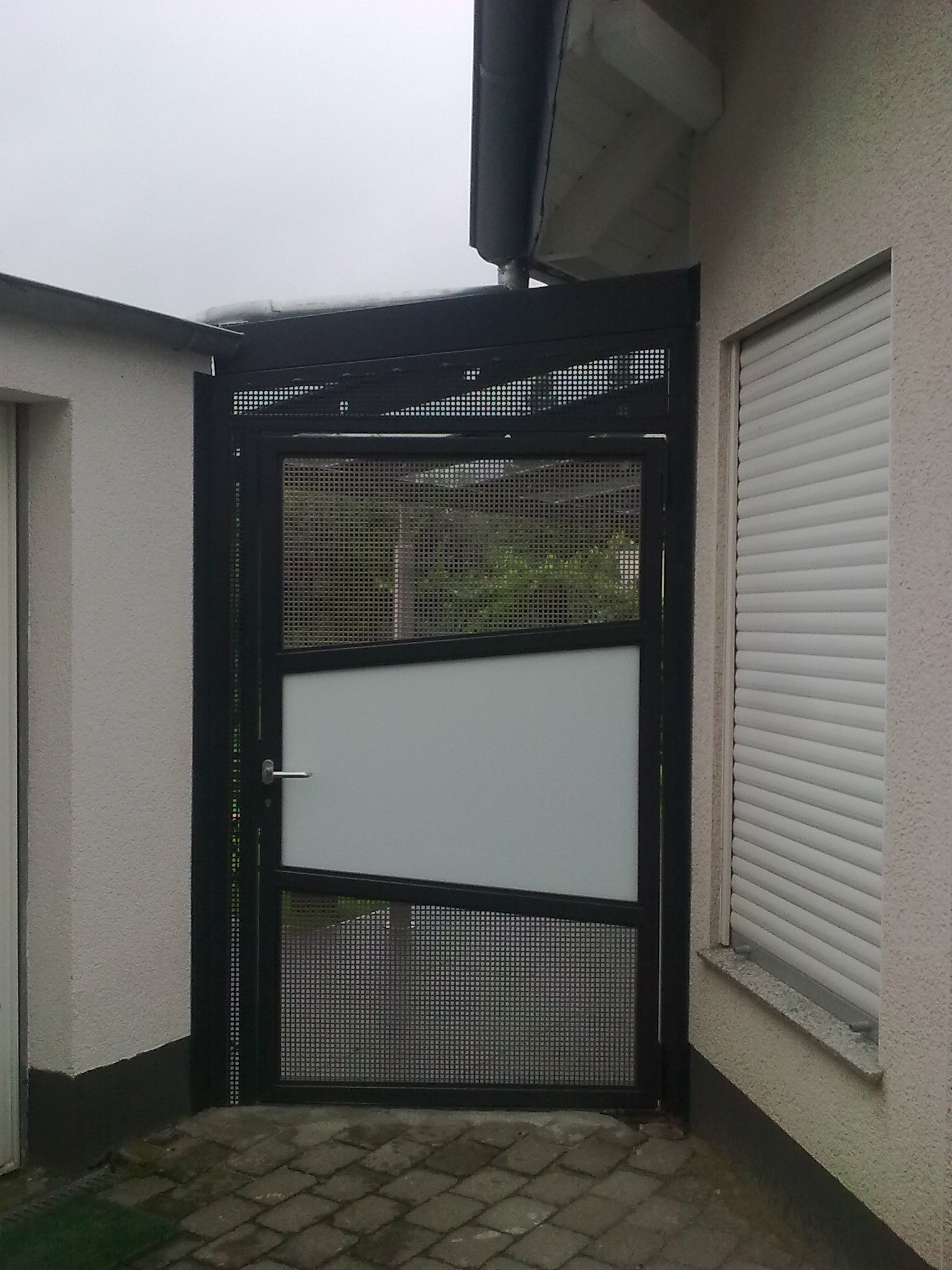 Gartentor aus schwarzem Stahl mit Gitterfüllung und Milchglasfeld im Durchgang zwischen zwei Gebäuden, darüber ein Gitter-Oberlicht bis unters Dach.