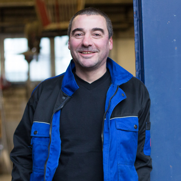 Thomas Jungbluth, Metallbauer, lächelnd in blau-schwarzer Arbeitsjacke neben einer blau beschichteten Stahlstütze, dahinter eine Werkhalle.