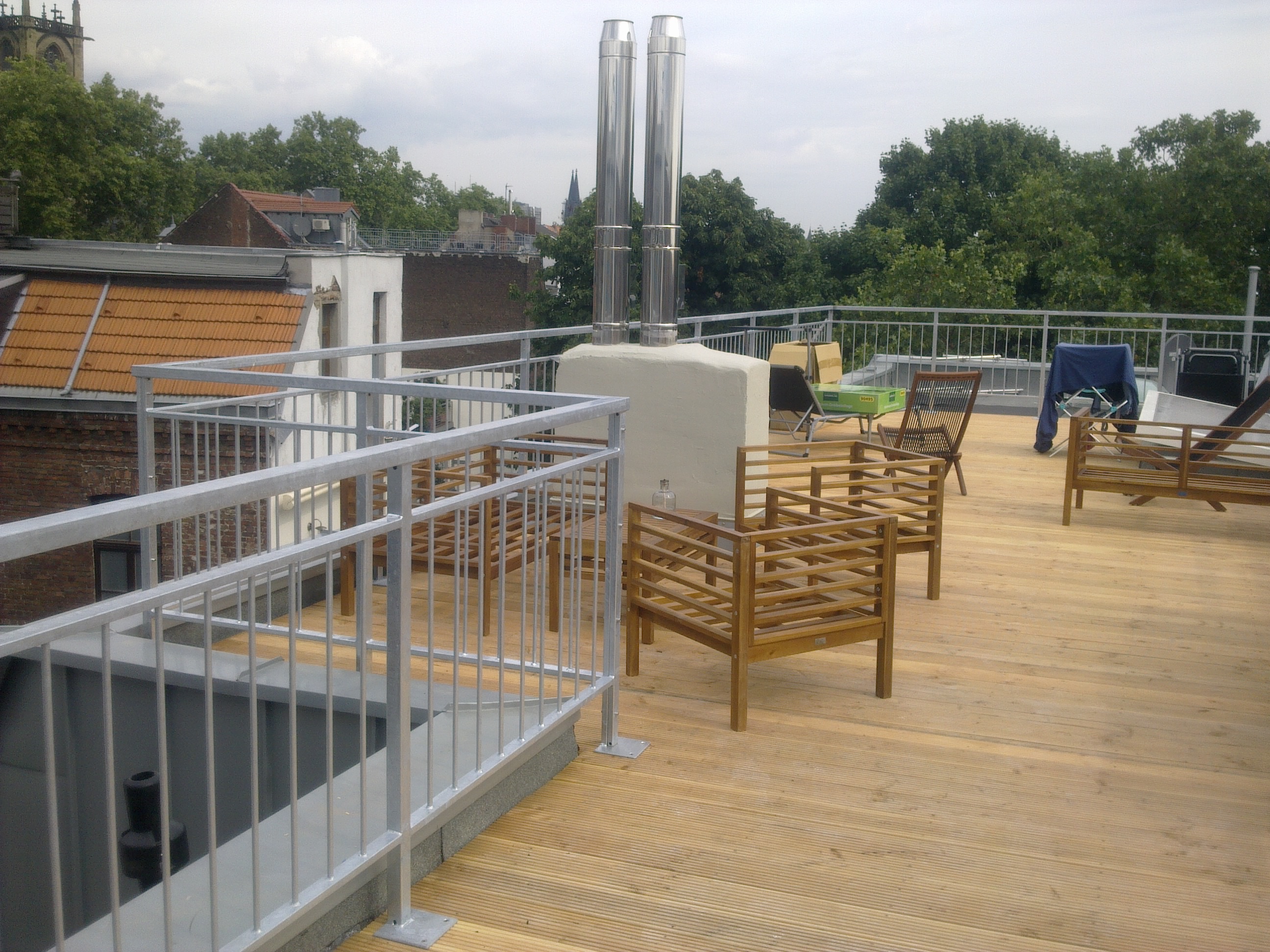 Verzinktes Geländer einer Dachterrasse mit senkrechten Stäben und Rohr-Handlauf über Eck, auf Holzdielen, dahinter die Dächer der Stadt.