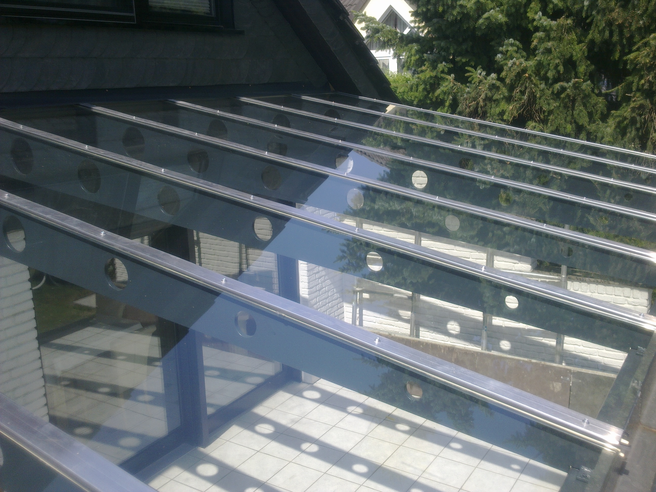 Terrassenüberdachung aus Glas auf beschichteten Stahlträgern mit runden Ausschnitten, schräg von oben, mit Schattenwurf auf den Fliesen.