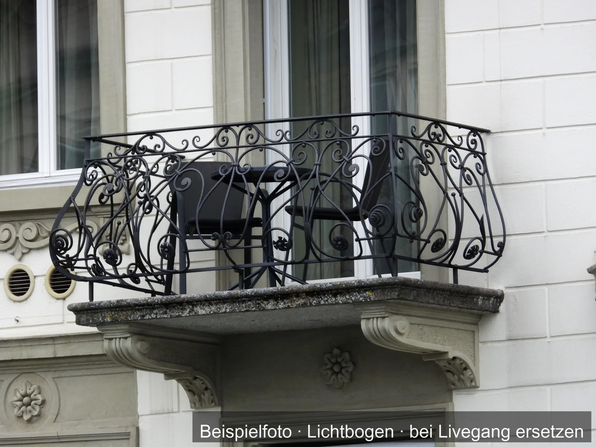 Kleiner Balkon mit geschmiedetem, verschnörkeltem Ziergeländer auf einer Steinplatte mit Konsolen an einer hellen Altbaufassade.