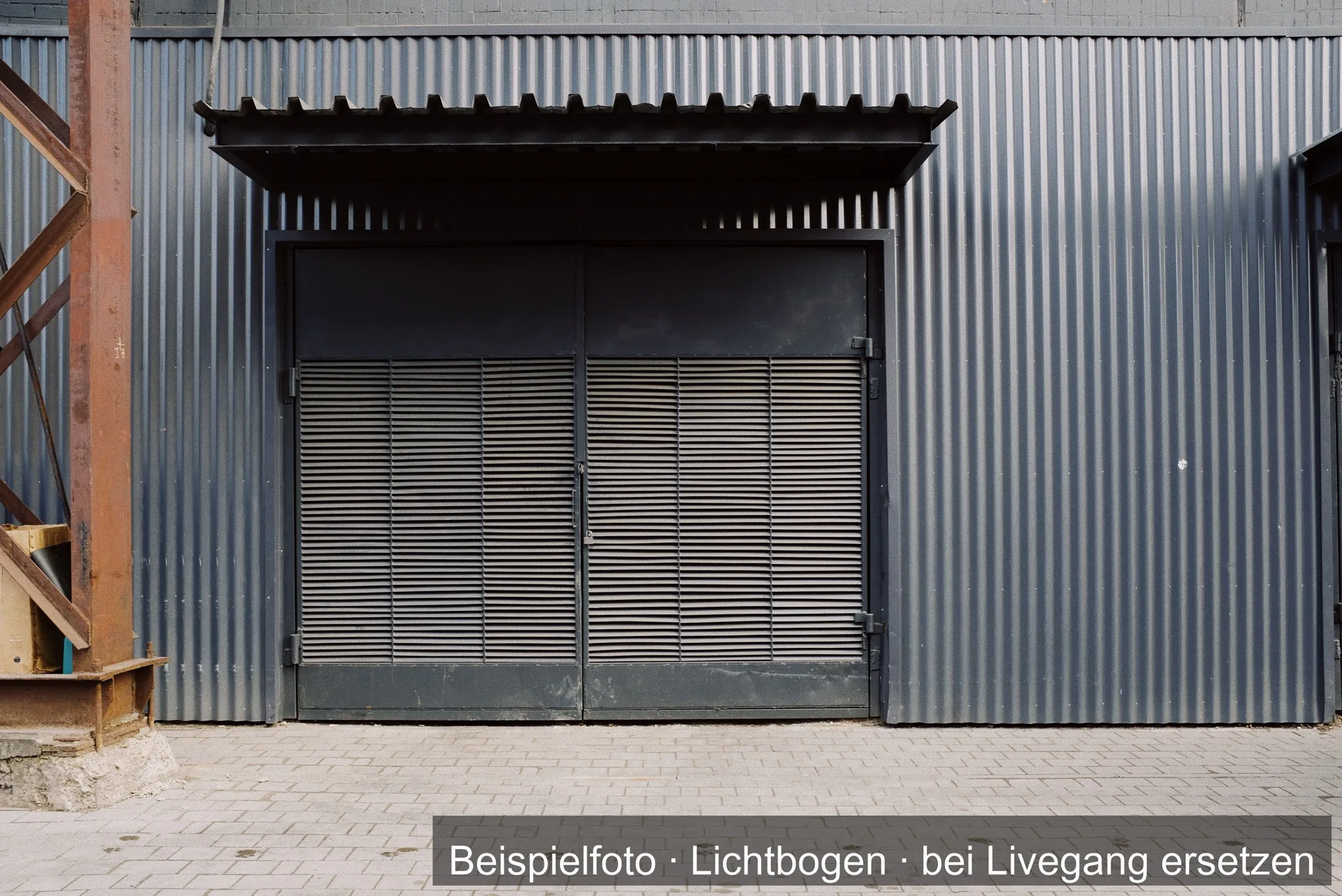Doppelflügelige Stahltür mit Lüftungslamellen unter einem kleinen Vordach in der grauen Wellblechfassade einer Halle.