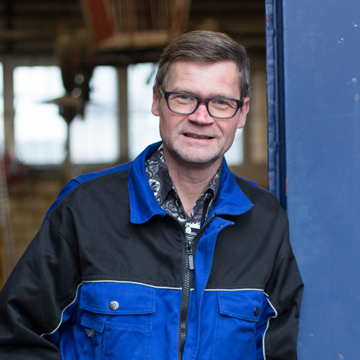 Michael Oberbörsch, Metallbaumeister, mit Brille in blau-schwarzer Arbeitsjacke neben einer blau beschichteten Stahlstütze, dahinter eine Werkhalle.